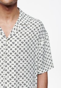 Camicia a maniche corte in tessuto bianco con motivo floreale e geometrico nero, colletto e bottoni frontali.