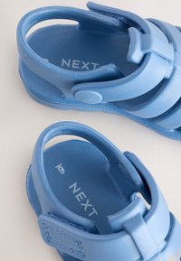 Sandales en plastique bleu avec un design à brides croisées, fermetures réglables à Velcro et intérieur texturé avec détail de logo de marque.