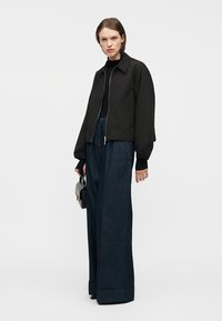 Γυναίκα με μαύρο μπουφάν με φερμουάρ, μαύρο τοπ, φαρδιά σκούρα τζιν παντελόνια, κρατώντας μια μικρή μαύρη τσάντα, στέκεται μπροστά σε απλό φωτεινό φόντο.