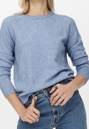 Vrouw draagt een lichtblauwe trui en een blauwe spijkerbroek, terwijl ze met beide handen de riemlussen van de spijkerbroek aanpast tegen een effen witte achtergrond.