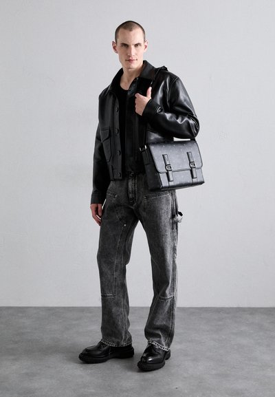 Giacca di pelle nera indossata sopra una camicia nera, abbinata a jeans in denim grigio e scarpe con suola spessa nere. Una borsa a tracolla nera con texture e cinghie.