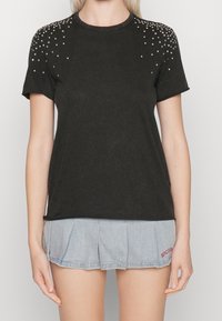 Schwarzes Baumwoll-T-Shirt mit kurzen Ärmeln, rundem Ausschnitt und dekorativen silbernen Nieten auf den Schultern. Kombiniert mit einem Jeansrock.