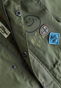 Chaqueta verde con cremallera y botones negros, decorada con parches que incluyen un cactus, una cara sonriente, texto "HAPPY SAFE," y "stay cool".