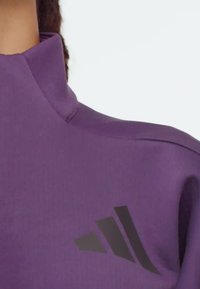 Fialový športový top s vysokým golierom a čiernym logom Adidas na hrudi, ktorý nosí osoba s tmavými vrkočmi.