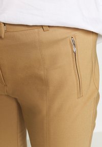 Beżowe spodnie wykonane z gładkiego materiału o dopasowanym kroju. Posiadają przednią kieszeń na zamek z srebrnymi detalami oraz przeszycia wzdłuż szwów.