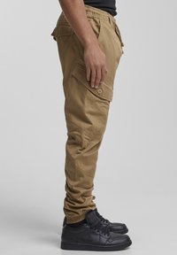 Tovor hlače v kaki barvi z elastičnim pasom, več žepi in sproščenim krojem. Kombinirane s črnimi supergami z nizkim profilom.