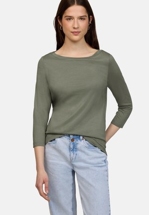 Ung kvinde iført olivengrøn top med trekvartærmer og lyseblå jeans, stående med den ene hånd i lommen mod en hvid baggrund.