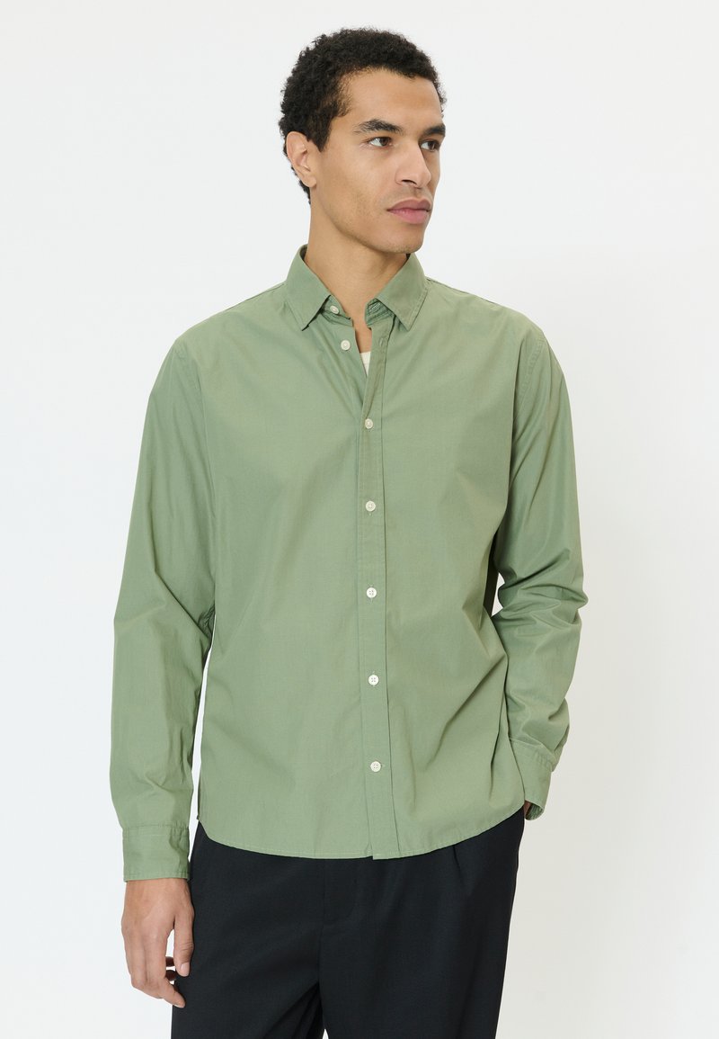 Homme portant une chemise à boutons vert clair avec un pantalon noir, regardant sur le côté devant un fond blanc uni.