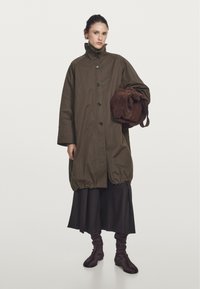 Przesadnie duży oliwkowy płaszcz z wysokim kołnierzem i czarnymi guzikami, w połączeniu z brązową zamszową torbą. Czarna spódnica i brązowe botki za kostkę dopełniają stylizację.