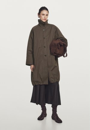 Übergroßer olivgrüner Mantel mit hohem Kragen und schwarzen Knöpfen, kombiniert mit einer braunen Wildledertasche. Ein schwarzer Rock und braune Stiefeletten runden den Look ab.