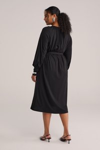 Schwarzes Midikleid mit langen Ärmeln, tailliertem Bund und glatter Textur. Mit abgerundetem Ausschnitt und dezentem Volumen an den Manschetten.