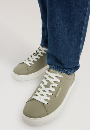 Persona che indossa scarpe da ginnastica verdi oliva chiaro con lacci bianchi e suole bianche, abbinate a jeans denim blu scuro.