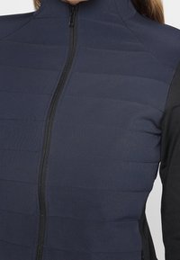 Veste hybride bleu marine avec un design matelassé structuré, dotée d'une fermeture éclair intégrale et de manches accentuées noires. Tissu lisse et léger.