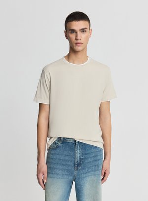 Giovane uomo con i capelli corti che indossa una maglietta beige sopra una canottiera bianca e jeans azzurri, in piedi su uno sfondo semplice.