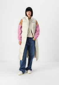 Gevoerde mouwloze jas in crèmekleur met kleurrijke pastel schouderaccenten; gecombineerd met een beige trui en blauwe wijde jeans.