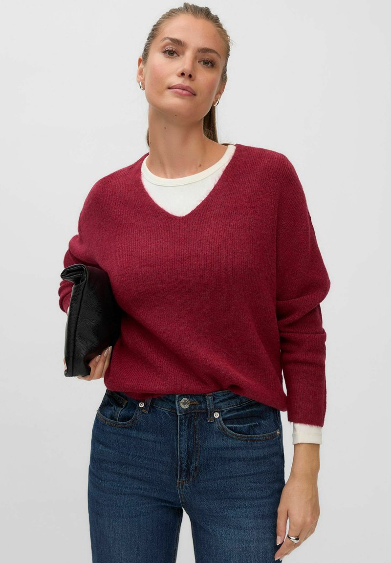 Femme portant un pull rouge à col en V par-dessus une chemise blanche et un jean bleu, tenant une pochette noire sous le bras.