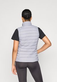 Šviesiai pilkas pūkinis liemenė su aukštu apykakle, vertikaliu siuvinėtu raštu ir prigludusia forma, derinama su juodu marškinėliu ir tamsiomis leggingais.