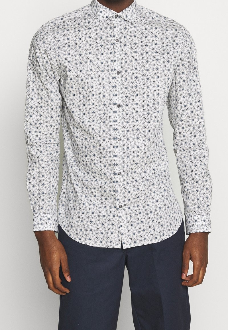 Biała koszula z długim rękawem z okrągłym wzorem w granatowym kolorze. Klasyczny kołnierzyk i gładki materiał. Noszona z ciemnymi spodniami.