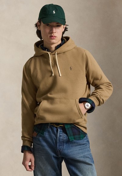 Jeune homme portant un sweat à capuche beige, une casquette verte, une chemise à carreaux, un jean bleu et une montre-bracelet, debout avec une main dans la poche.