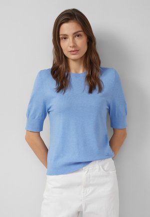 Frau mit langen braunen Haaren trägt einen hellblauen Kurzarm-Pullover und weiße Hose, steht vor einem schlichten grauen Hintergrund.