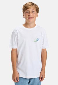 Bílý bavlněný tričko s barevným grafickým designem na levé hrudi. Krátké rukávy a kulatý výstřih. Kombinováno se světle modrými džíny.