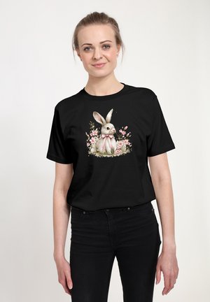 Žena v černém tričku s potištěným designem bílého králíka s růžovou mašlí sedícího mezi růžovými květinami.