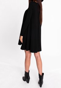 Abito nero a maniche lunghe con silhouette svasata, realizzato in tessuto morbido, indossato con stivaletti neri alla caviglia, visto da dietro.