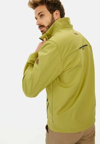 Lysegrøn softshell-jakke med høj krave, lynlås foran og lommer, med et læderlogo på ærmet og sorte detaljer.