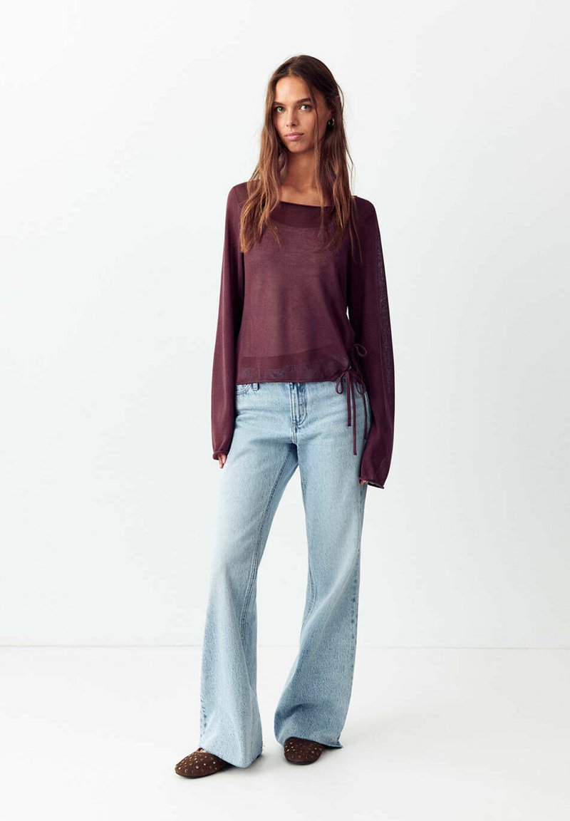 Femme debout portant un haut transparent bordeaux à manches longues avec des liens sur les côtés, un jean large bleu clair et des chaussures marron sans lacets.