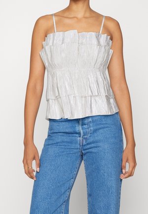 Vrouw draagt een zilveren geplooid mouwloos topje met ruches en blauwe spijkerbroek, staand tegen een effen achtergrond.