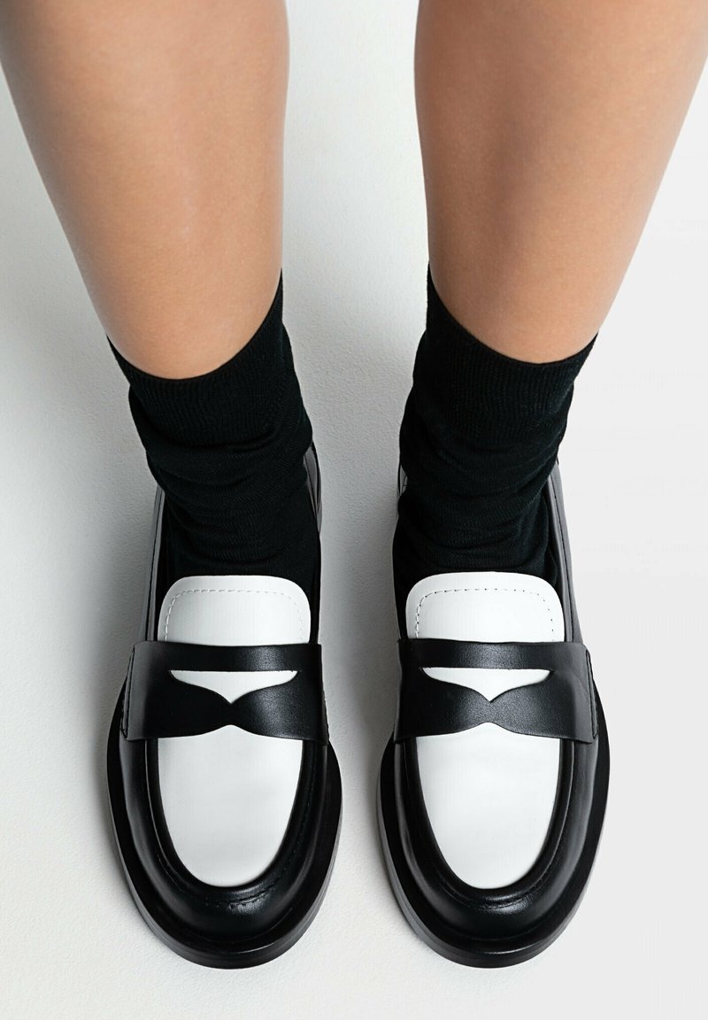 Pieds portant des mocassins noir et blanc avec des chaussettes noires sur un fond clair, vus de dessus.