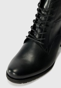 Bottine en cuir noir avec une finition lisse, bout rond et fermeture à lacets. Elle dispose d'une semelle en caoutchouc basse et d'un détail de couture subtil.