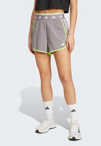 Naine kannab halli Adidas'i lühikesed püksid, millel on ere roheline äär, valgeid Adidas'e sokke ja valgeid jooksujalatseid, seistes heledal taustal.