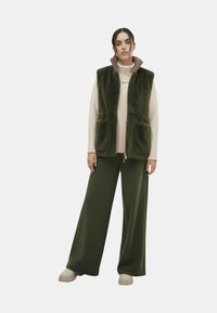 Groen fleecevest met twee voorzakken, gedragen over een crème coltrui. Een paar olijfgroene broek met wijde pijpen en lichtgekleurde schoenen.