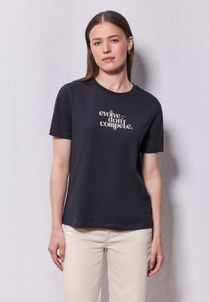 T-shirt en coton noir à manches courtes et col rond. Présente un texte stylisé blanc qui dit "évoluer-ne pas rivaliser."
