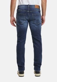 Vista trasera de una persona que lleva vaqueros de mezclilla azul oscuro, camiseta blanca y zapatillas negras y blancas, de pie con los brazos relajados a los lados.