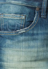 Los jeans de mezclilla de lavado claro presentan un color azul desgastado, acentos destruidos, un bolsillo estándar y costuras en marrón a lo largo de las uniones.