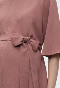 Κοντινό πλάνο υφάσματος φορέματος σε χρώμα σκόνης τριαντάφυλλου με ζώνη που δένει σε φιόγκο στη μέση και φαρδιά μανίκια μήκους έως τον αγκώνα σε άτομο.