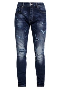 Jeans de denim azul escuro com um corte slim, apresentando áreas desgastadas, bordas desfiadas e manchas de cor clara espalhadas para um efeito texturizado.