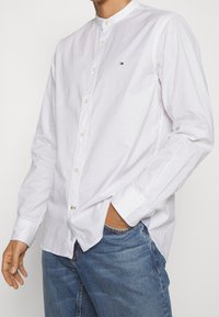 Camicia bianca con colletto alla cinese, con accenti blu a contrasto e un piccolo logo sul petto, abbinata a jeans di denim blu.