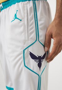 White basketball shorts with teal accents, featuring mesh fabric, side pockets, and a large black hornet logo on the thigh.