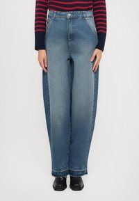 Jeans in denim azzurro chiaro a gamba larga con vita alta, caratterizzati da una chiusura a bottoni, tasche laterali e orlo sfrangiato. Indossati con scarpe nere.