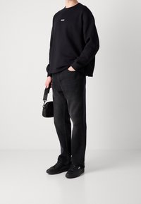 Persona vistiendo sudadera negra con el logo "HUGO", jeans negros, zapatillas negras, sosteniendo una pequeña bolsa negra en la mano contra un fondo blanco.
