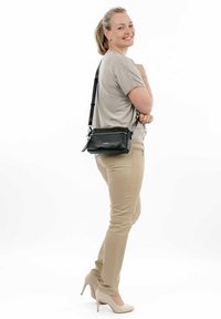 Borsa a tracolla in pelle nera con chiusura a zip, hardware argentato e tracolla regolabile, indossata da una persona con pantaloni beige e tacchi.