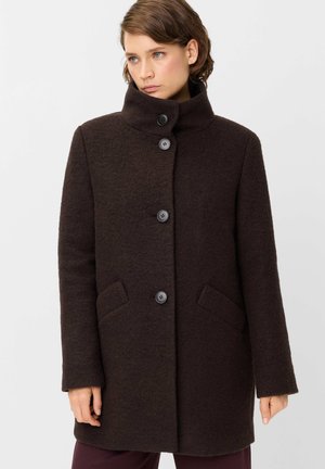 Woman with short brown hair wearing a dark brown buttoned coat with high collar and her hands relaxed by her sides.