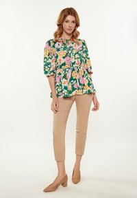 Blusa floreale verde con maniche a sbuffo e un fiocco al collo, abbinata a pantaloni attillati beige e tacchi color tan.
