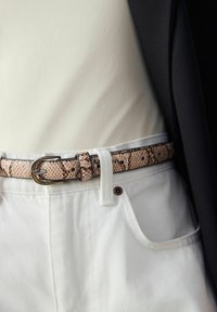 Ceinture à motif de peau de serpent en brun clair avec une boucle métallique, portée sur un haut crème et un pantalon blanc, présentant une finition texturée.