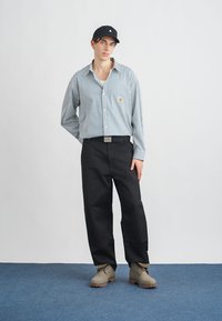 Camisa de manga larga a rayas, de color azul claro y blanco, con un parche de logo; pantalones anchos negros; botas grises; gorra negra. Diseño simple y moderno.