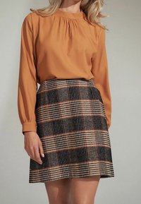 Blusa a maniche lunghe color senape con scollo plissettato abbinata a una gonna a quadri nei colori nero, beige e arancione, dal taglio strutturato.