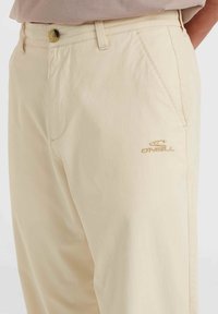 Pantalons beiges en tissu léger, offrant une coupe classique, des poches latérales et un logo brodé sur la jambe.